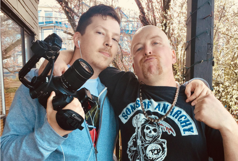 Two caucasian men embrace each other for a photograph. The one on the left wields a camera while the other, on the right, raises his fist.