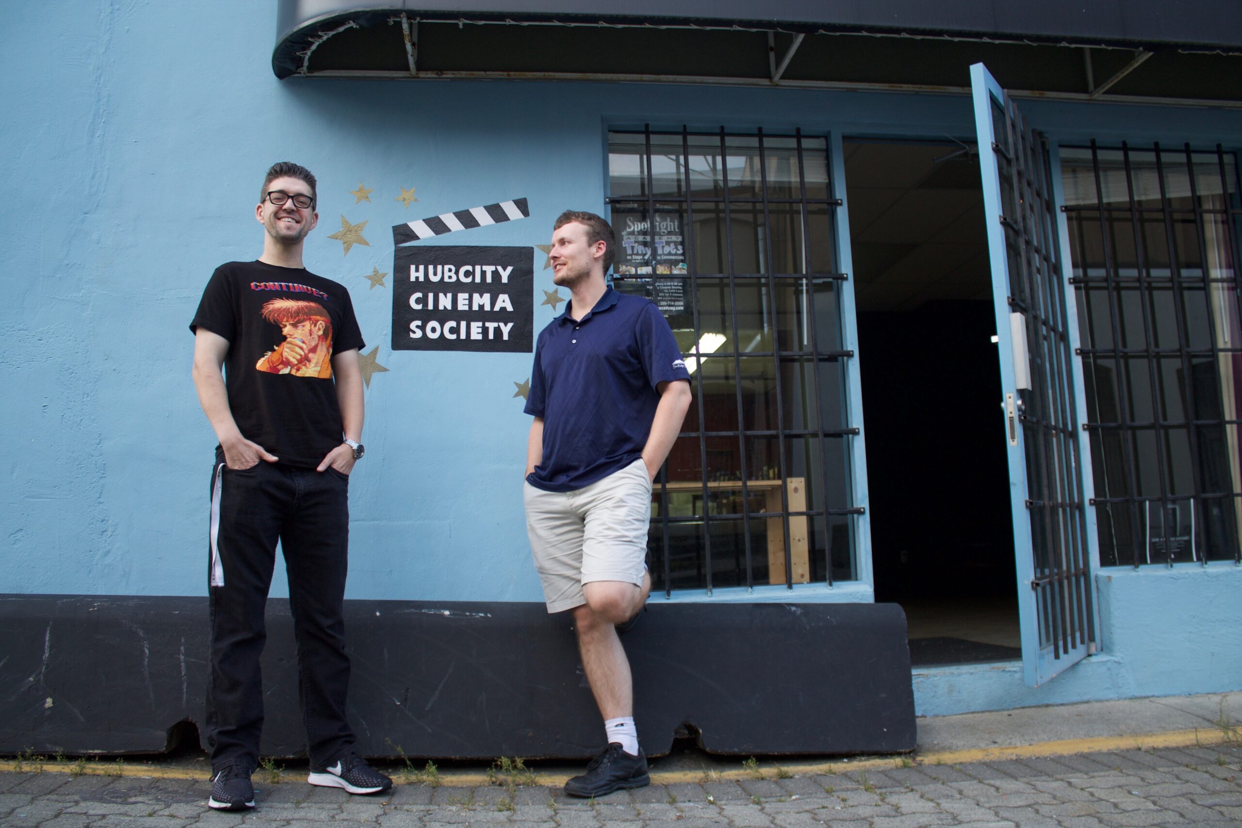 Two men stand a meter apart at the entrance of HCCS’s headquarters. The door to society is open while both men entertain each other; the logo of the nonprofit organization is painted on the wall and between both co-founders. The logo is a clapperboard with the name Hub City Cinema Society within it.