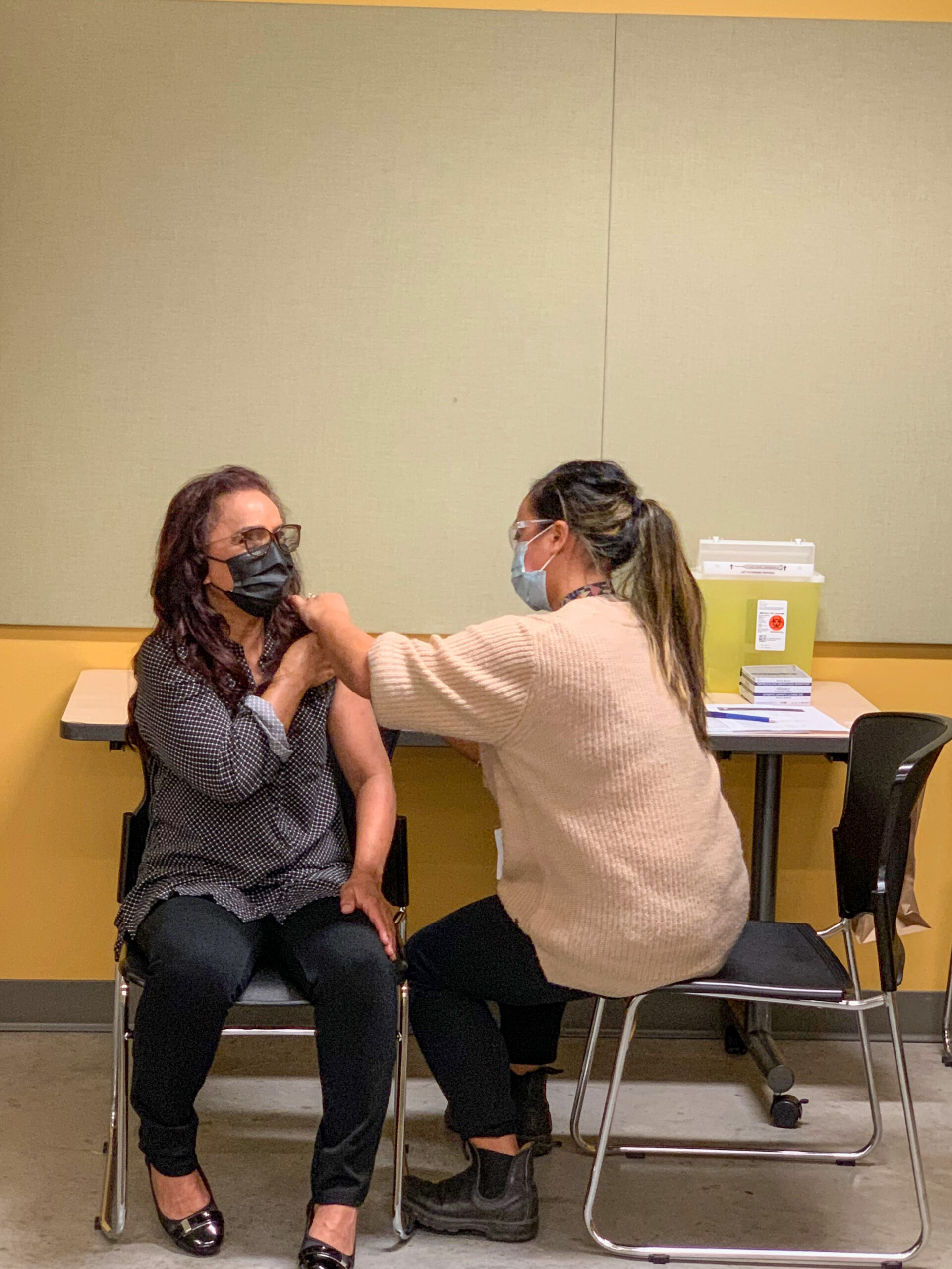 Pictured is Nisha Yunus sitting down on a chair wearing a black face mask as she receives her immunization. The nurse beside her is wearing a non-medical blue face mask as she pokes the needle into Nisha's arm.